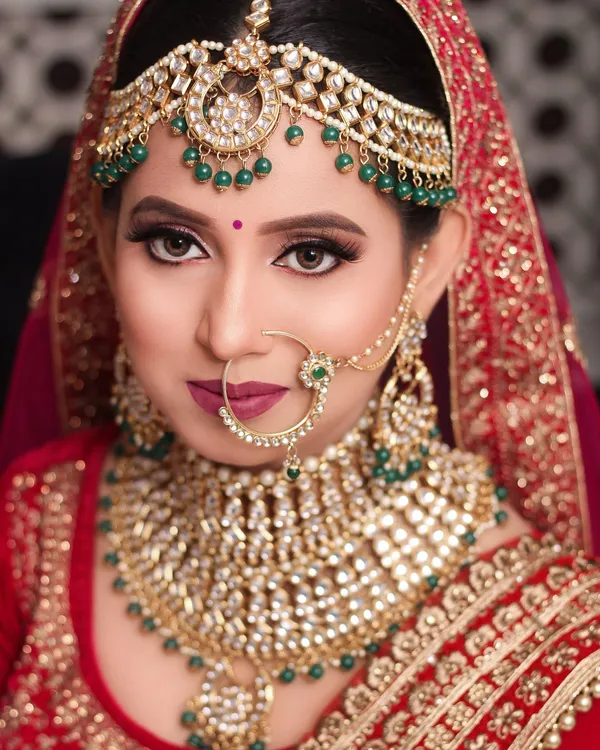 Bride in red with emerald and kundan jewellery and a gold maang tikka, bridal makeup by Blushed By Aashi in Lucknow